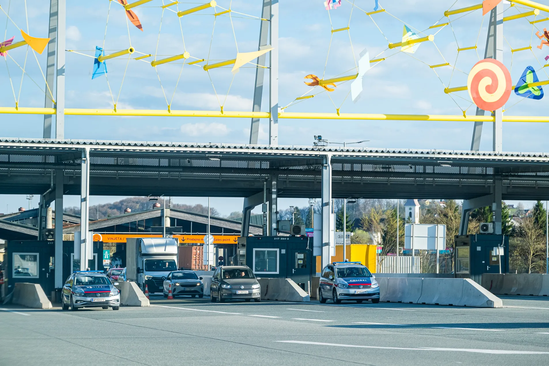 Slovenia–Austria Šentilj border crossing with Austrian Polizei VW patrol cars, orange Vinjeta sales point gantry and colourful artistic installation overhead, April 2026
